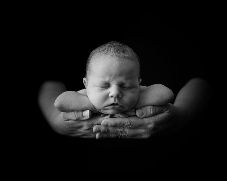 black and white photo of a newborn baby laying on her chest and hand in the palm of her daddy hands.