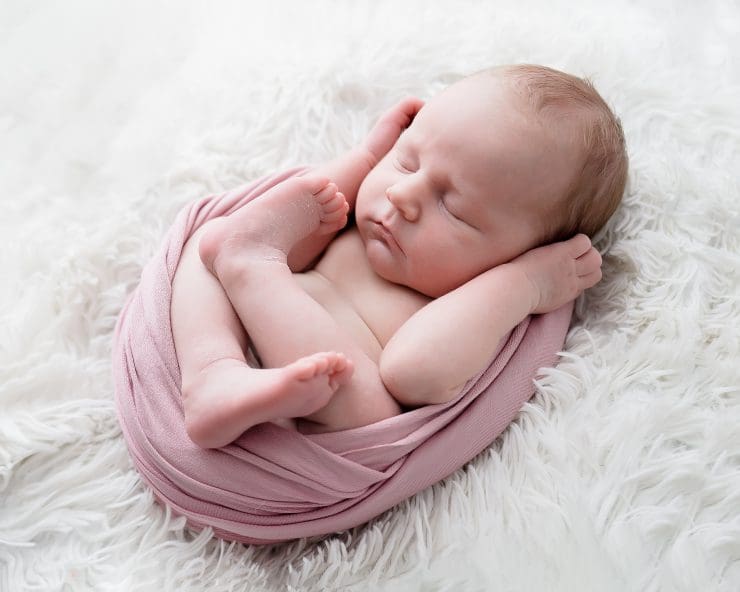 baby ada all wrapped up in a pink fabric and laying on a white fluffy rug with her arms up