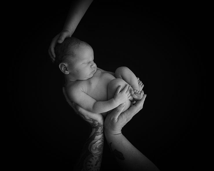 Black and White Newborn baby photo, laying on a black fabric and being held with mum, dad and big sisters hands