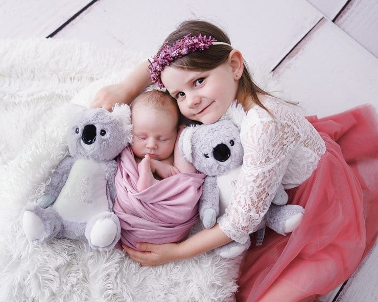 newborn baby wraped up in a pink fabric while big sisters lays at the side of her with teddies.
