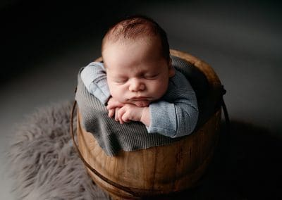 baby boy newborn photo where the baby is in a wooden bucket prop and leaning on his hands fast asleep