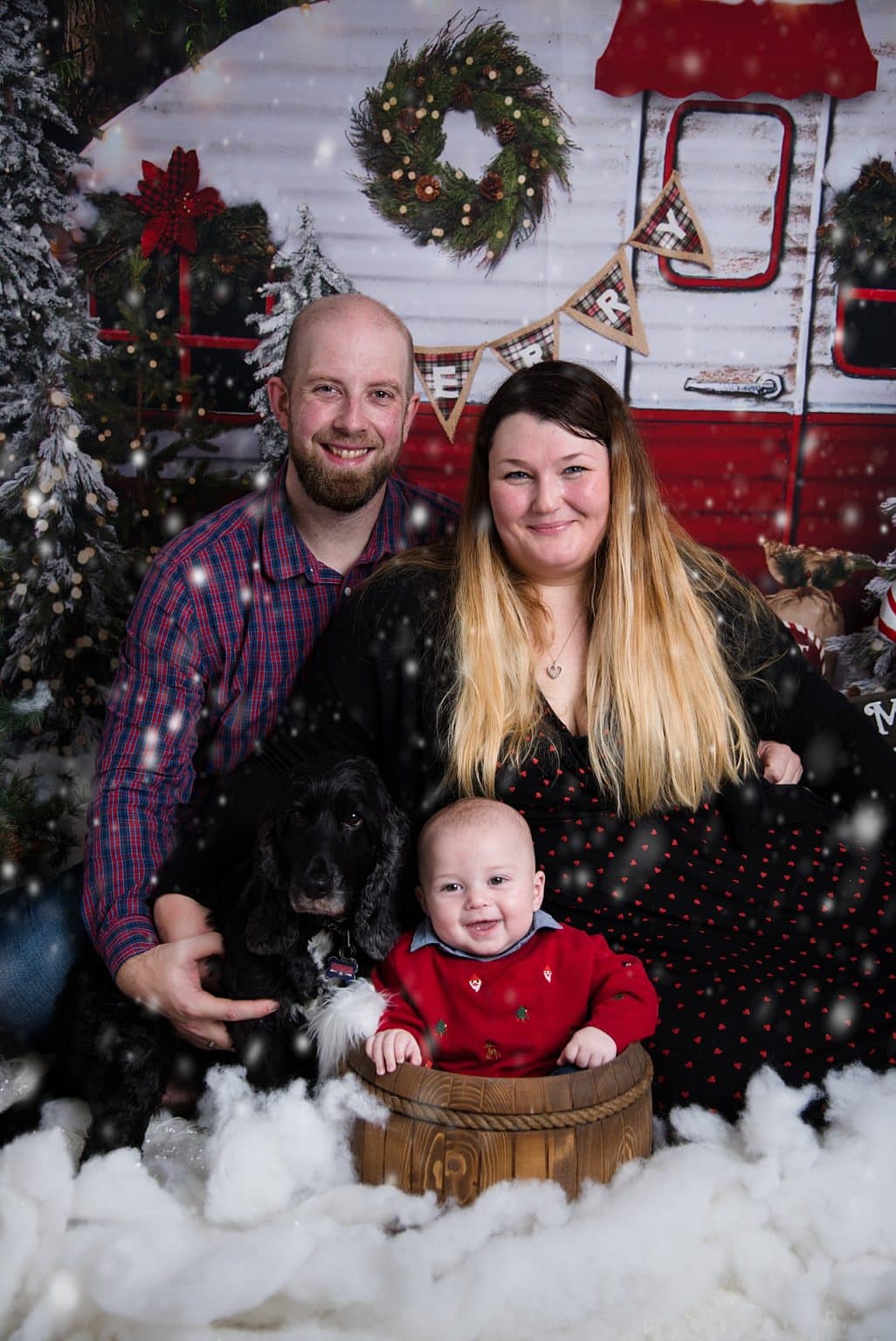 A family poses with a dog and baby in a festive, snowy scene. The man, woman, and baby are from left to right, with the baby sitting in a wooden barrel. A red and white caravan and decorated trees are in the background.