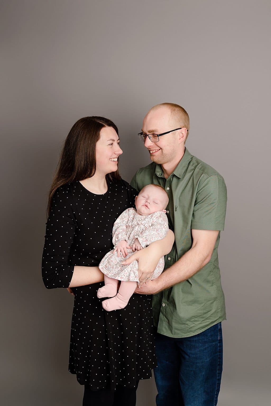 A smiling couple holds a sleeping baby. The woman has long brown hair and is wearing a black dress with white dots. The man is wearing glasses and a green short-sleeve shirt. The baby is dressed in a patterned outfit and pink socks. The background is grey.