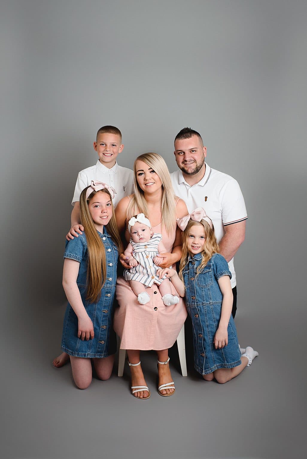 Family portrait with five people against a gray background. A woman in a pink dress holds a baby in the center, flanked by a man in a white shirt and three children: a boy and two girls in denim dresses, smiling at the camera.