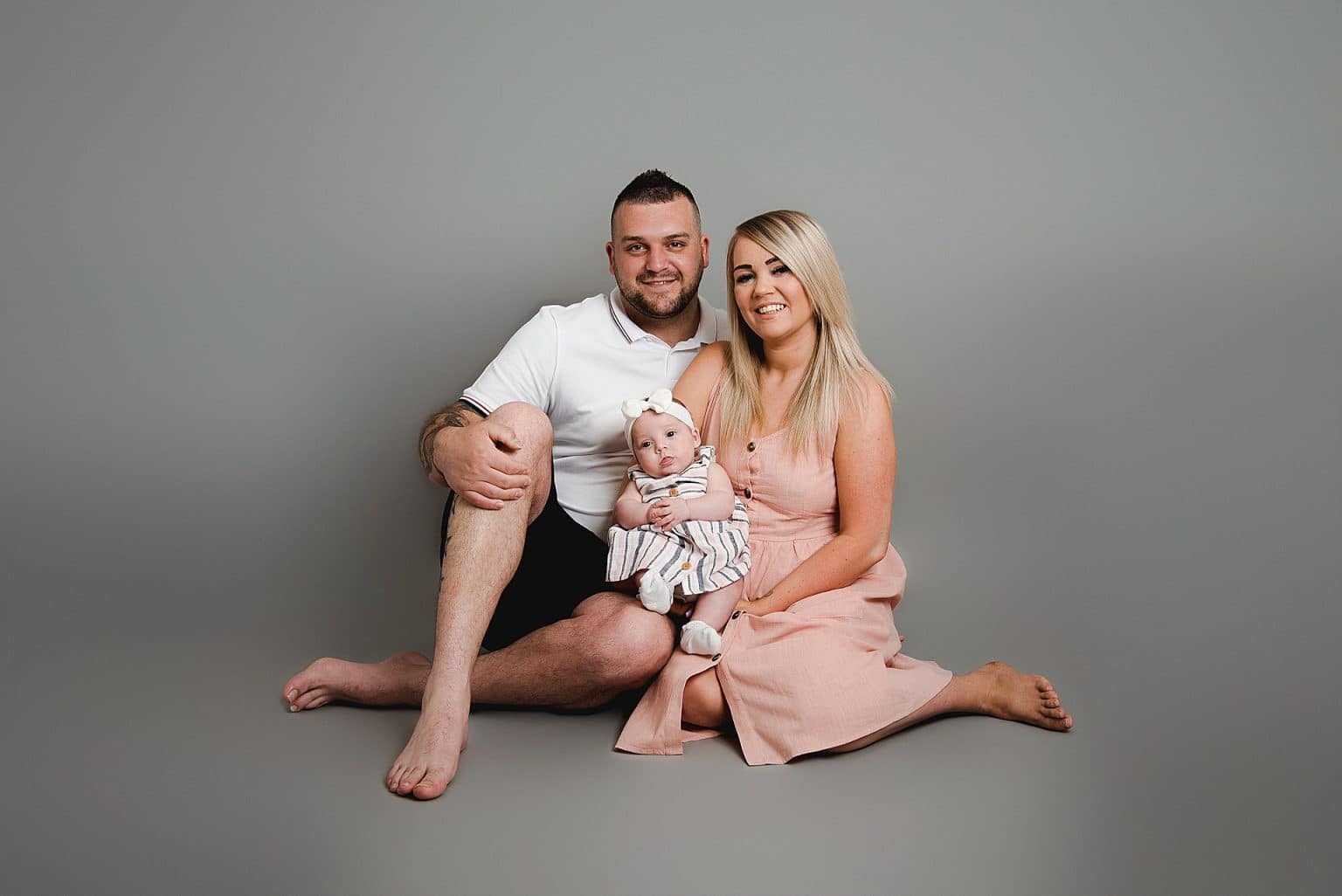 A couple sitting on the floor against a gray backdrop, smiling, with a baby in their arms. The man is wearing a white polo shirt and black shorts, while the woman is in a peach dress. The baby is in striped overalls with a headband.