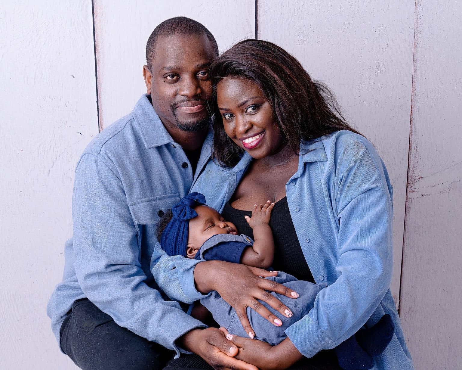 A smiling couple sits closely, each wearing a light blue shirt. The woman is holding a sleeping baby dressed in blue, with a matching headband. The background is a light, textured wall. They appear content and close-knit.