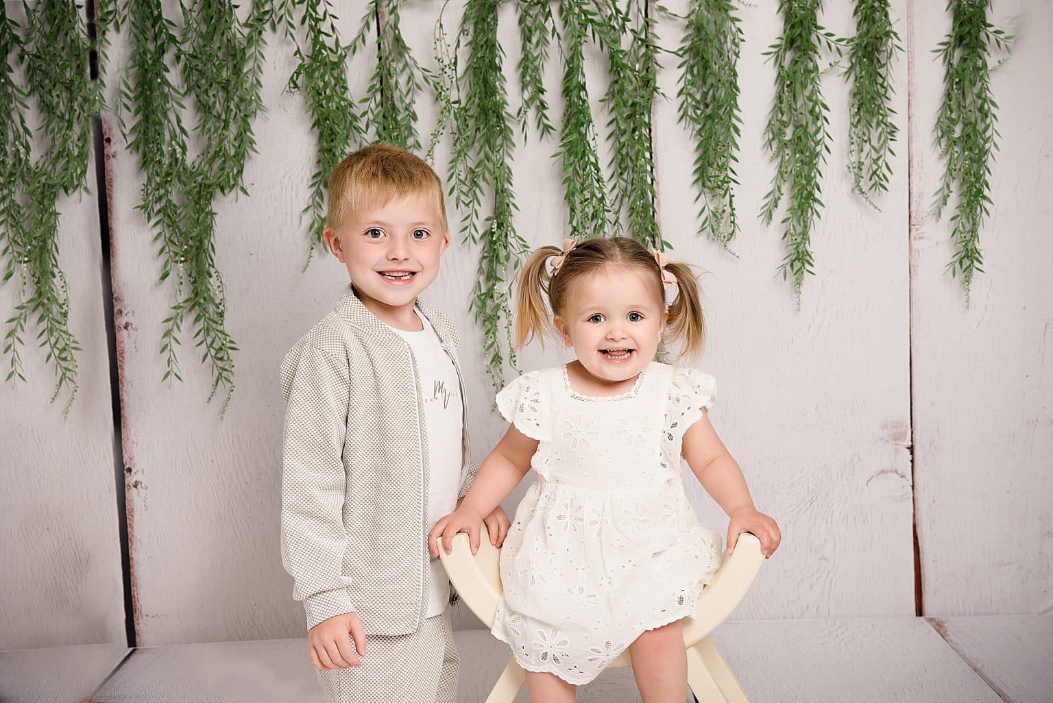 Two smiling children pose in front of a wooden wall with hanging greenery. The child on the left stands wearing a light-colored outfit, while the child on the right sits in a chair, wearing a white dress with pigtails.