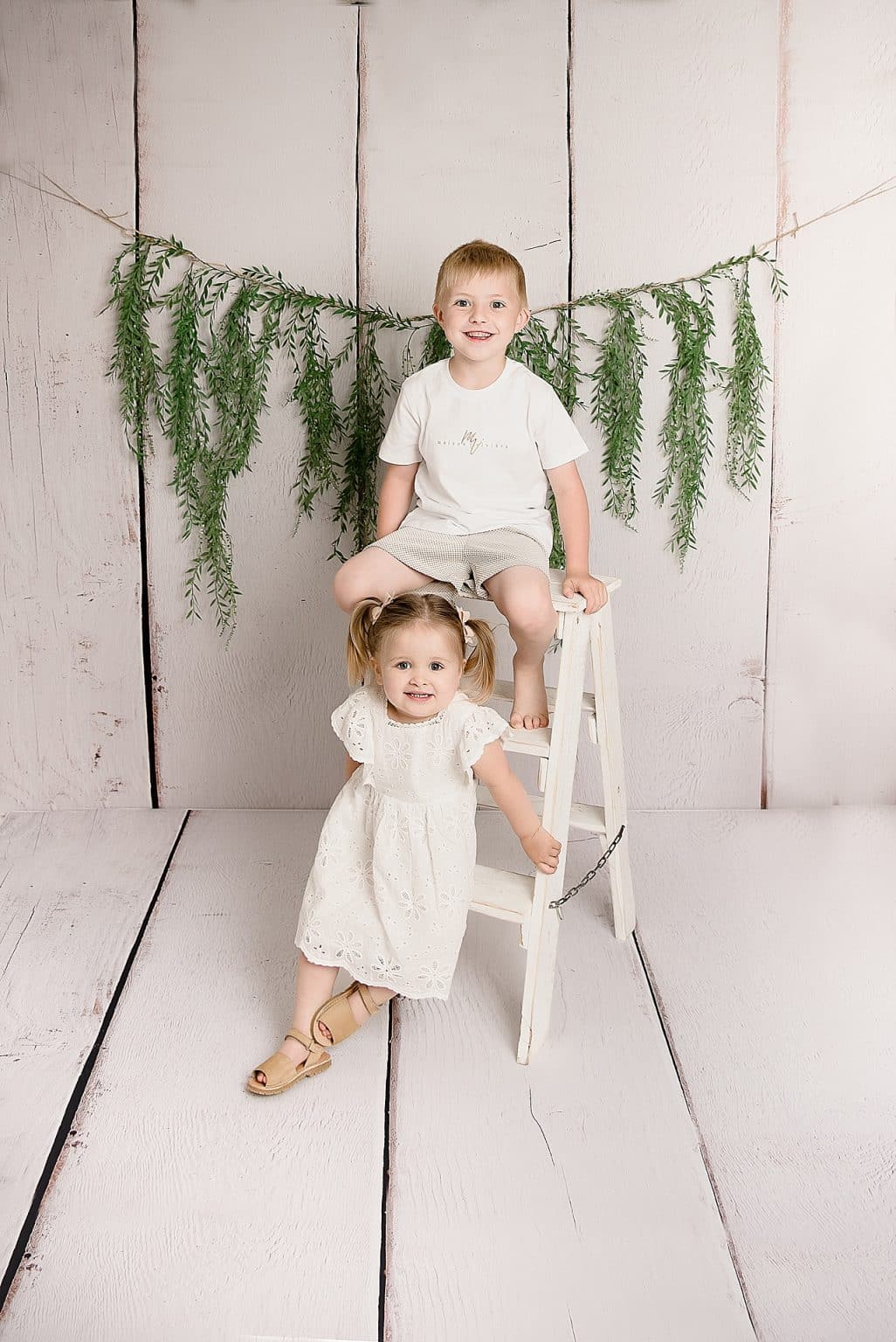 Two young children in a white, rustic setting. A boy sits atop a white ladder wearing a white shirt and shorts. A girl in a white dress stands beside the ladder. Greenery is hung in the background.