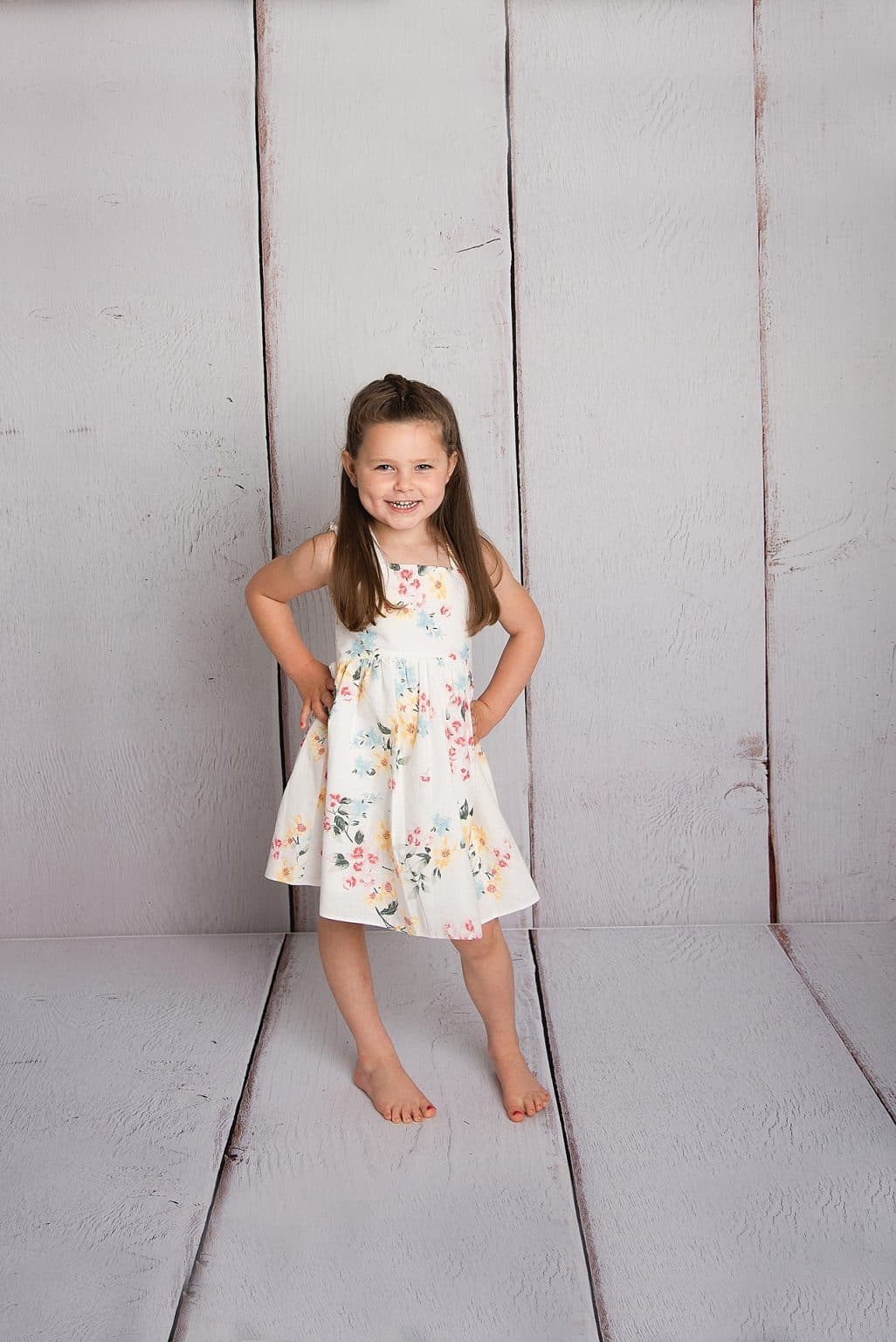 A young girl stands barefoot on a wooden floor, wearing a white dress with colorful floral patterns. Her long hair is down, and she has her hands on her hips, smiling at the camera against a light gray wood backdrop.