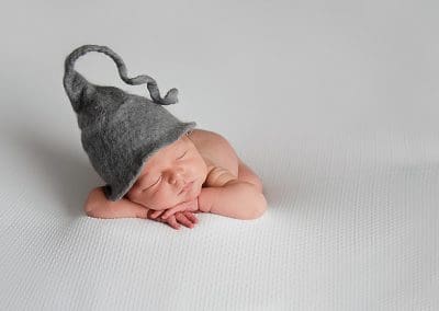 newborn baby fast asleep on his hands, laying on a white blanket with a grey pixie hat on