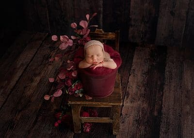 cute baby girl during her newborn photo shot, fast asleep in a red bucket and the bucket is on a chair with some flowers to the side