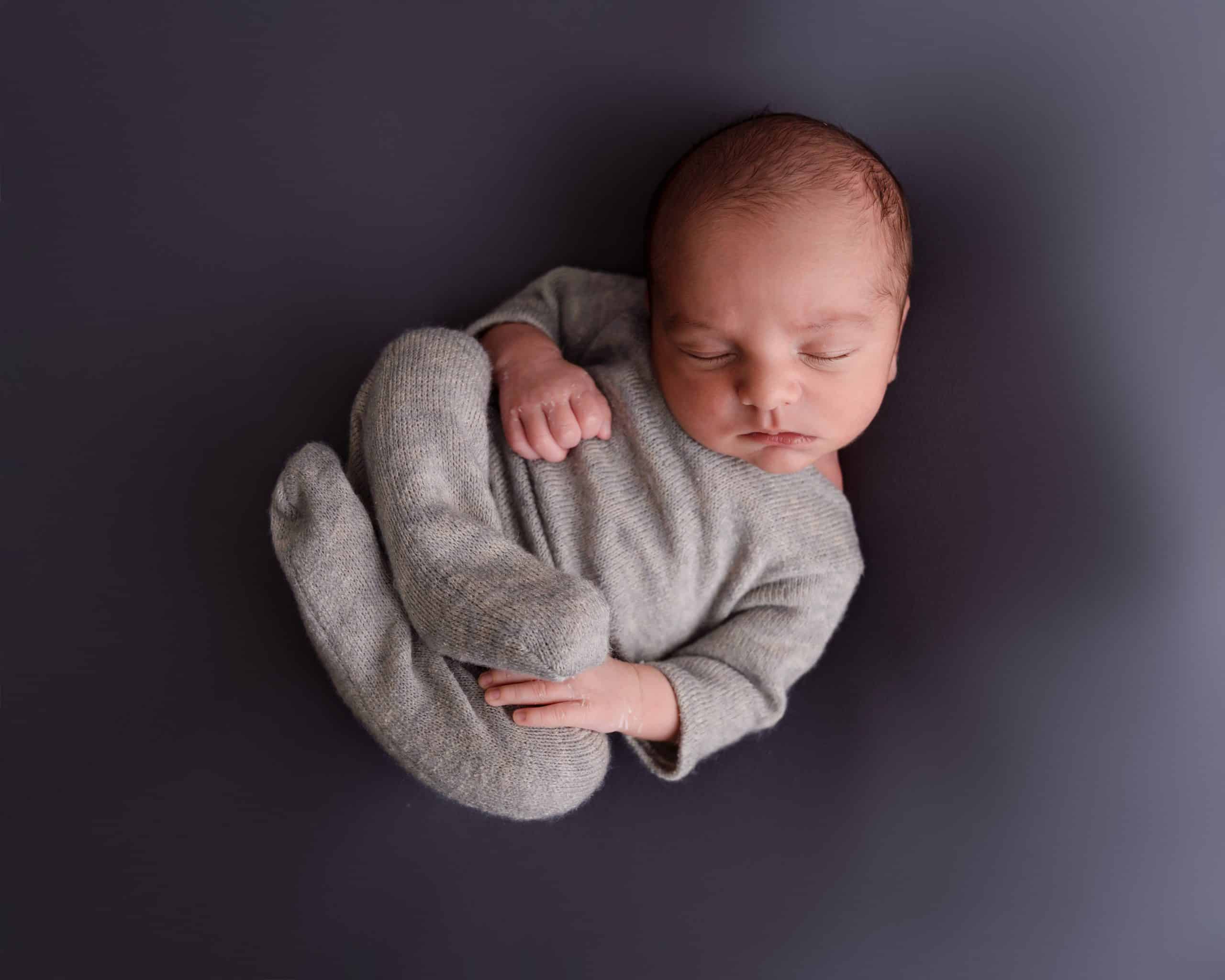 Newborn baby boy laying on his back all curled up in a grey outfit.he is on a grey backdrop fast asleep during his newborn session in Mansfield, Nottingham.