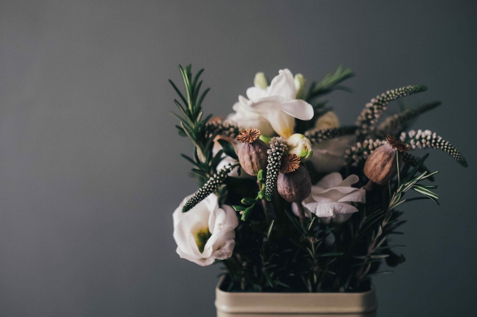 A bouquet in a vase against a gray background, featuring white flowers, dried seed pods, and sprigs of greenery.