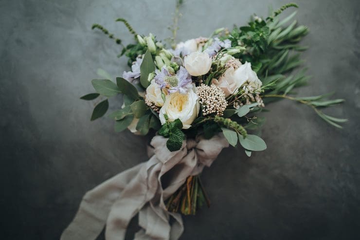 A bouquet of various flowers, including white roses, greenery, and delicate pastel blooms, is tied together with a light, flowing gray ribbon, set against a dark, textured background.