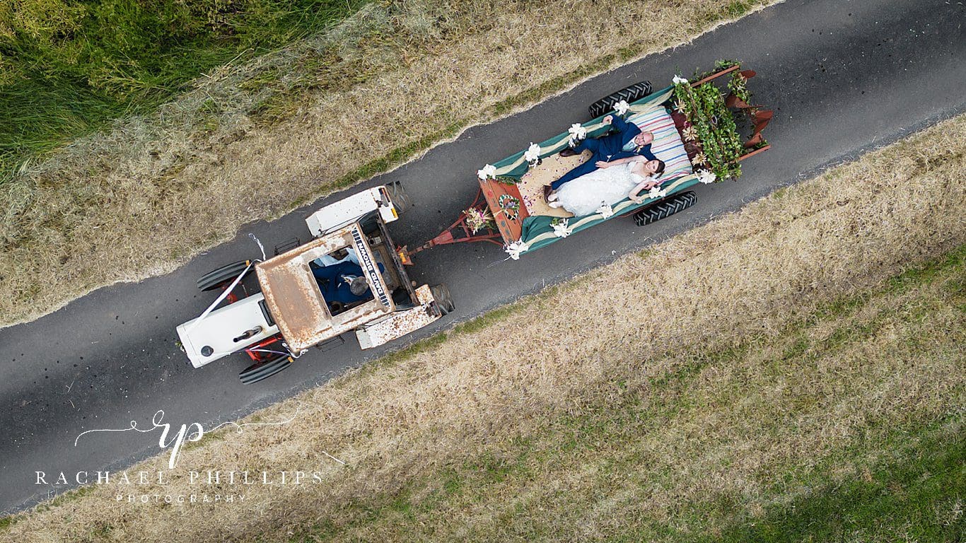 Drone photo of the bride and groom who are in the back of a trailer being pulled by the family tractor and the brides dad is the driver.