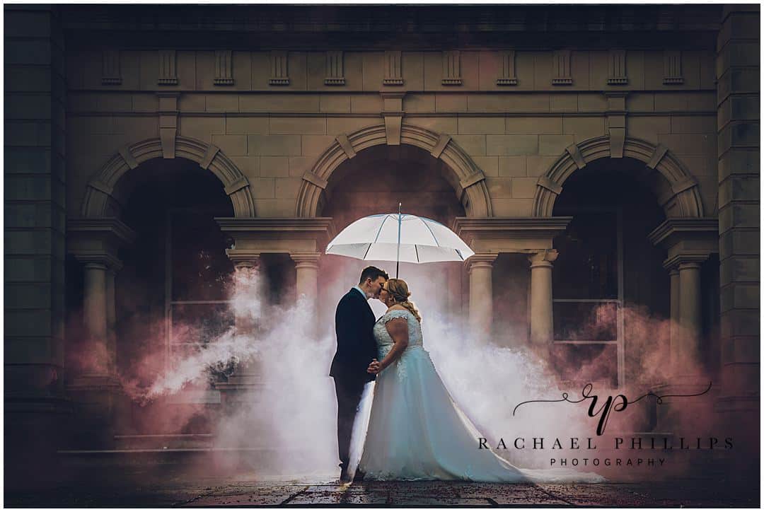 Couples photo in the rain with some added smoke from some Enola smoke bombs. The bride and groom are stood under an umbrella outside Alfreton Hall in derby