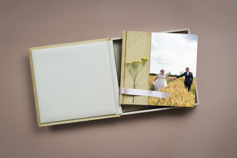 A wedding photo album lies open in a cardboard box. The cover features a bride and groom holding hands in a wheat field. Two yellow flowers and a white ribbon embellish the album.