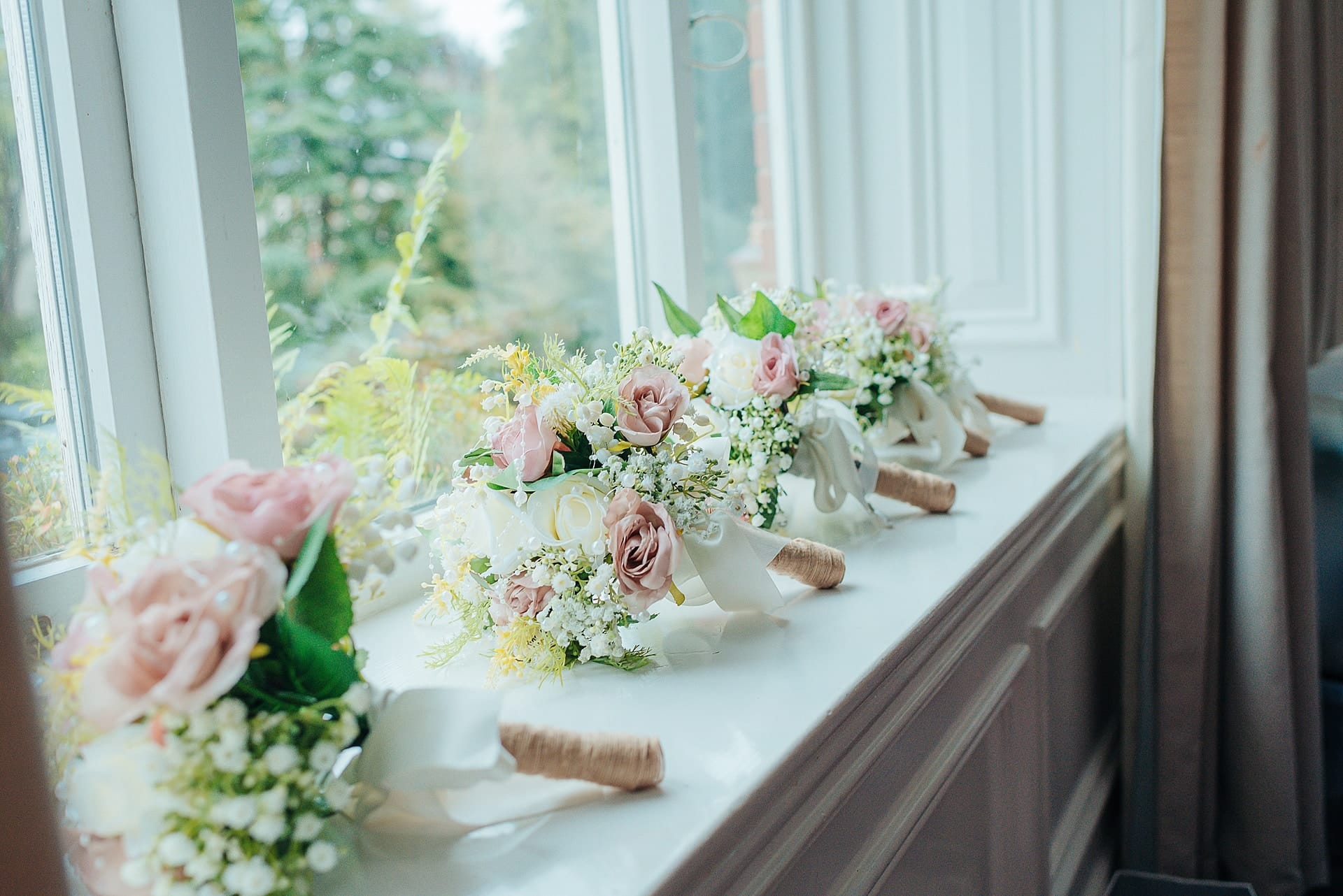 Bouquets of pastel pink and white flowers with greenery are lined up on a white windowsill. Sunlight filters through the window, illuminating the soft colors of the blossoms and elegant white ribbons attached to the bouquets.