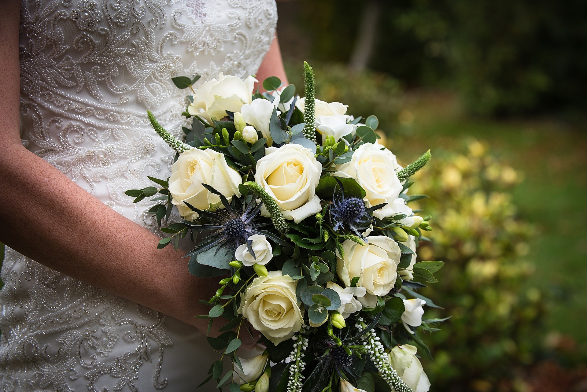 A bride in a white lace dress holding a cascading bouquet of white roses, greenery, and small blue thistles. The background is blurred greenery.