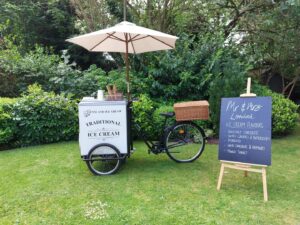 An ice cream bike with an umbrella is parked on grass next to a chalkboard sign. The sign lists ice cream flavors like "Belgian Chocolate" and "Mango Sorbet." Lush greenery surrounds the scene.