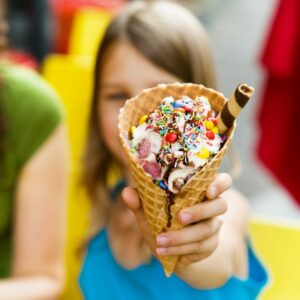 A person holds a large ice cream cone topped with colorful sprinkles, candies, and a chocolate wafer stick. The background is blurred, with hints of vibrant colors suggesting an outdoor setting.