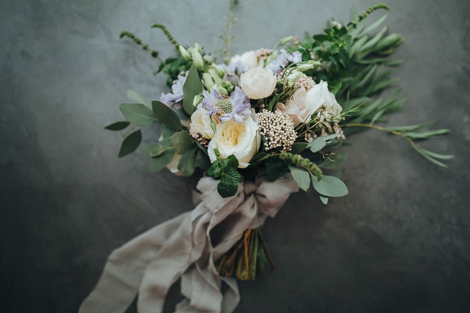 A bouquet of various flowers including white roses, lilac blossoms, and greenery, tied with a wide, flowing gray ribbon, rests on a textured gray background.