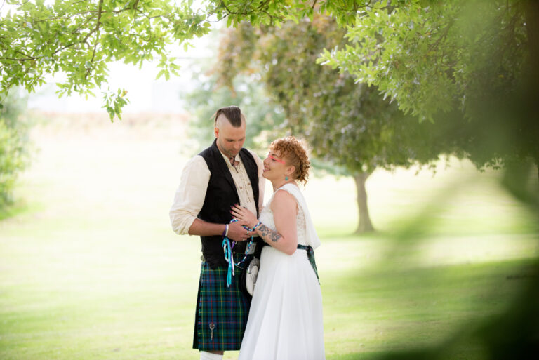 Couple in kilt and dress under trees, outdoors.