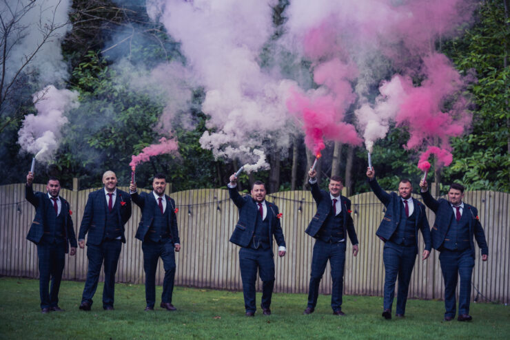 Groomsmen holding colourful smoke flares outdoors.