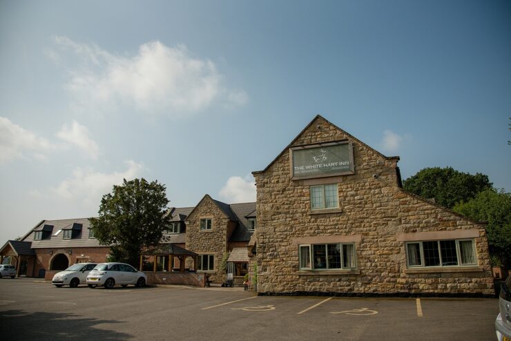 Stone building of "The White Hart Inn" under a blue sky with fluffy clouds. The structure features multiple windows and a parking area with a few cars. Trees flank the building, adding greenery to the scene.