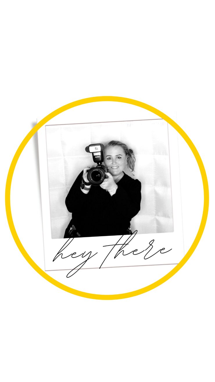 A black and white photo of a person smiling and holding a camera toward the viewer. The image is framed with a yellow circle and has the handwritten text "hey there" at the bottom.