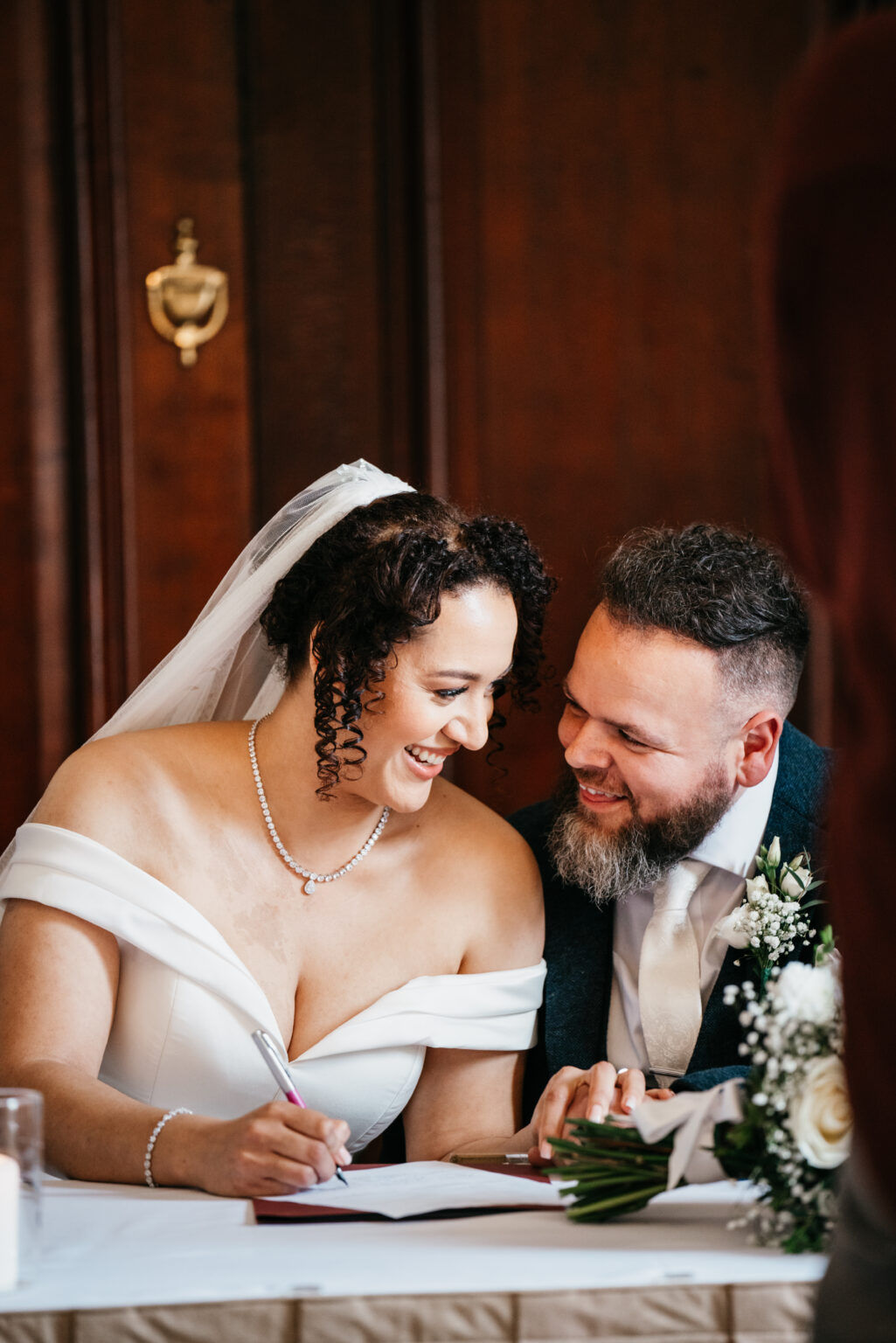 A bride and groom sit closely together. The bride, in an off-the-shoulder white dress, smiles while signing a document. The groom, wearing a dark suit, looks at her fondly. A bouquet of white flowers is on the table.