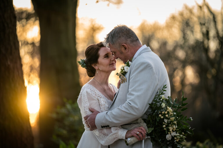 A couple embraces outdoors, wearing formal attire, with the sun setting behind them. The woman holds a bouquet and both have gentle expressions, leaning their foreheads together amidst a backdrop of trees.
