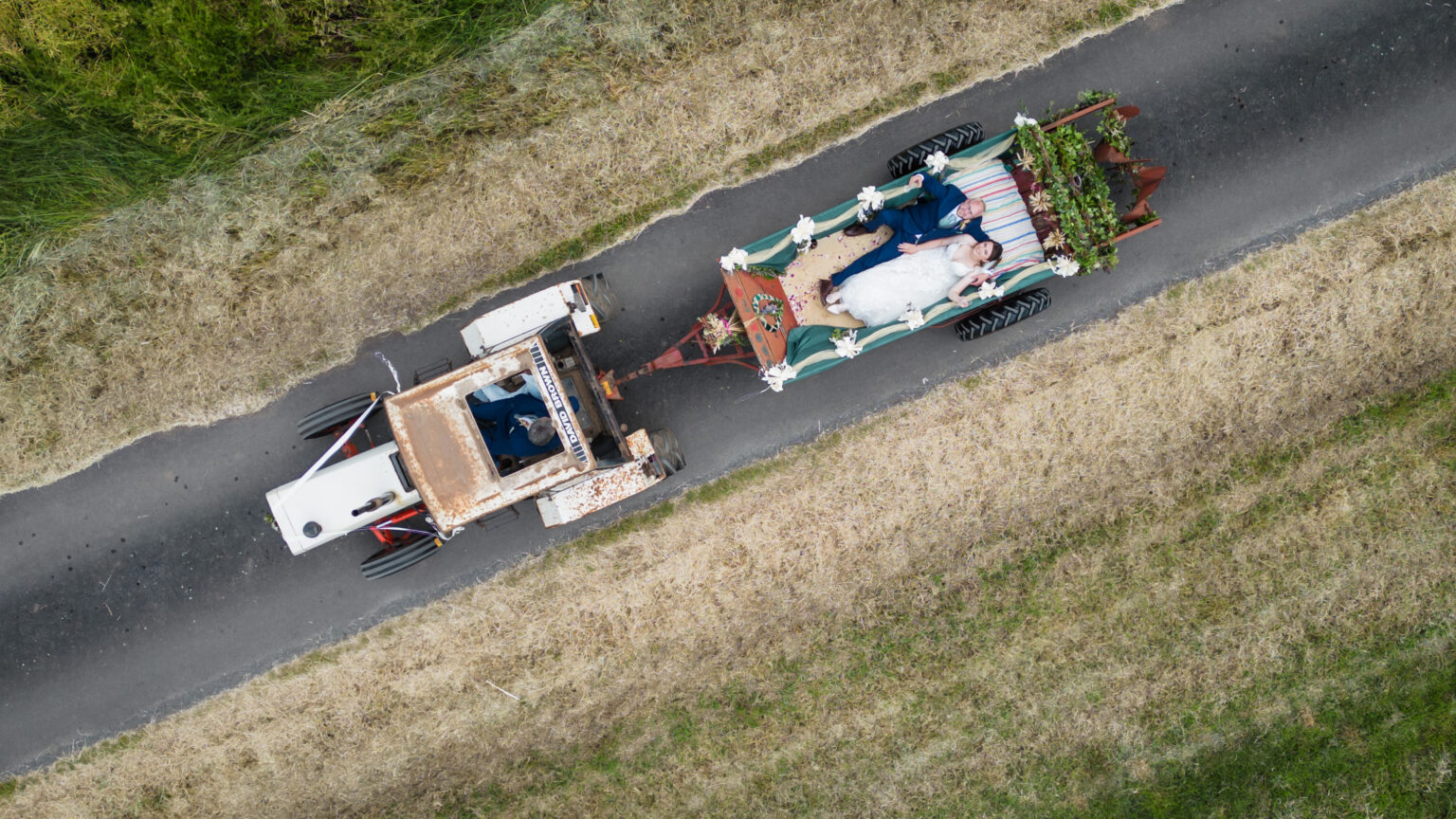 Aerial view of a rustic tractor pulling a decorative carriage on a countryside road. The carriage is adorned with flowers and carries two people dressed in wedding attire, surrounded by open grassy fields.