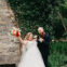 Bride in a white gown holds a bouquet and poses playfully with a man in a dark suit and red beard. They are standing in a garden with a stone wall covered in green ivy. Both look joyful and celebratory.