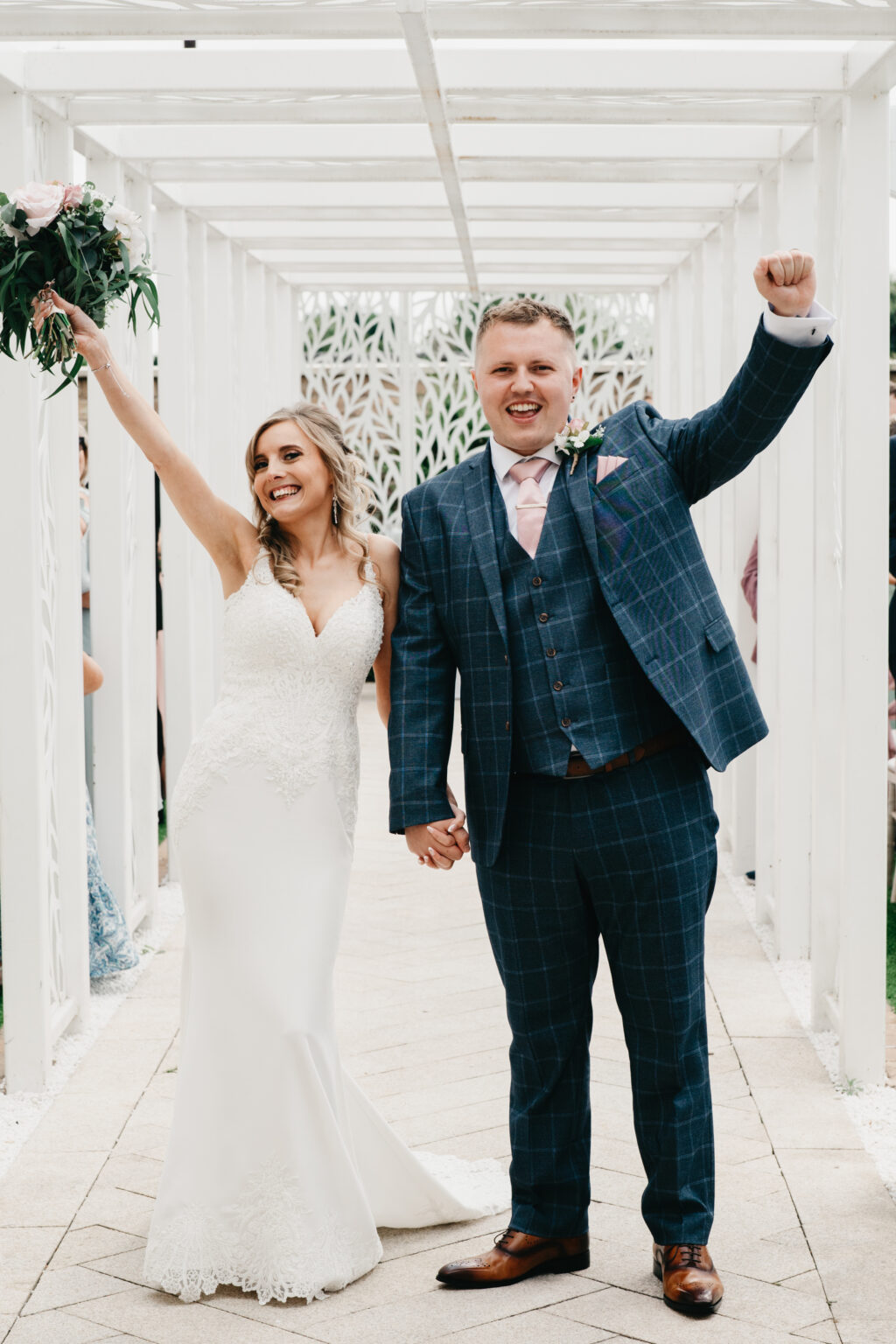 A happy bride in a white dress and a groom in a blue checkered suit joyfully raise their arms under a white pergola. They hold hands, and the bride holds a bouquet of flowers, celebrating their wedding.