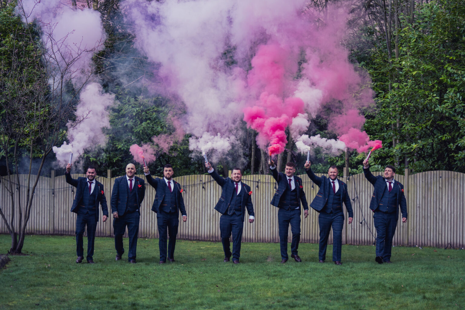 Six men in suits walk on grass holding smoke flares emitting pink and white smoke. They are in an outdoor garden setting with green trees and a wooden fence in the background.
