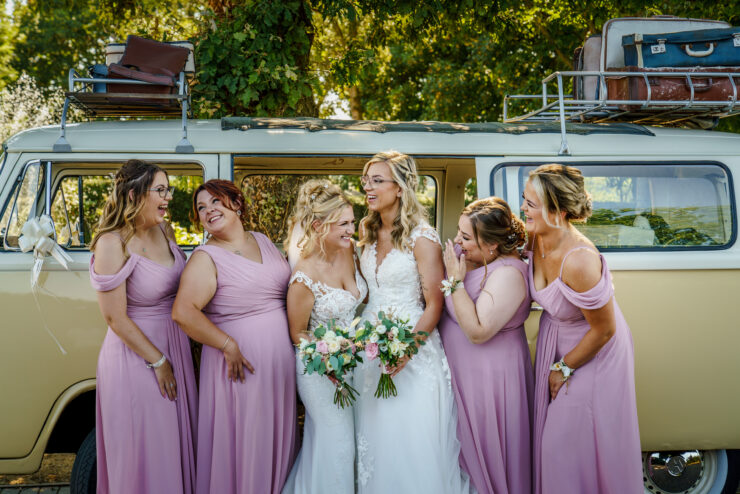 A bride and five bridesmaids in lavender dresses stand smiling in front of a vintage yellow van with luggage on top. The bride holds a bouquet of flowers. They are surrounded by greenery and sunlight.