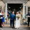 A smiling couple in wedding attire walks down steps with guests on either side throwing confetti. The groom holds up a fist in celebration, and the bride carries a bouquet. The setting is a building with columns.