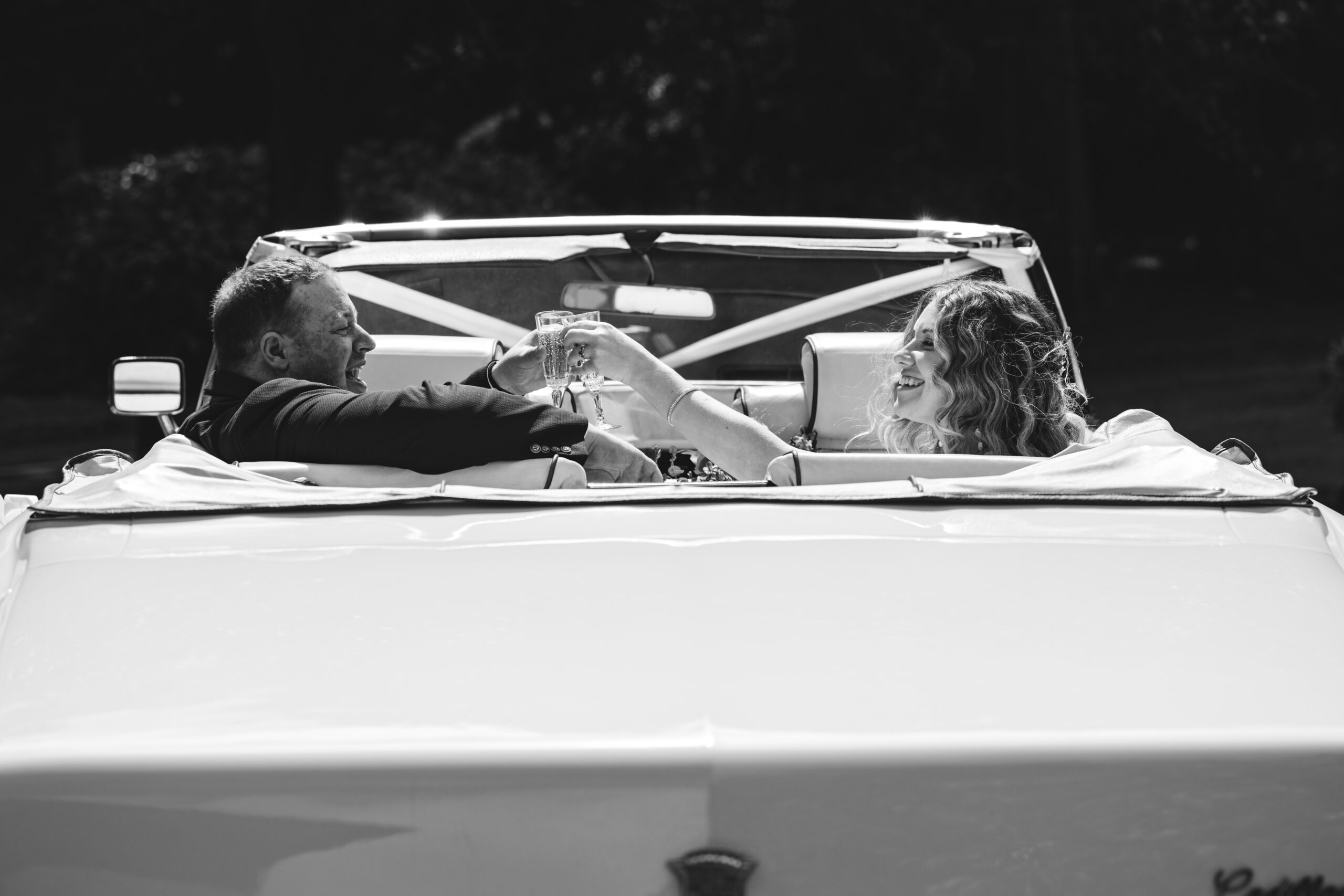 A black and white photo of a couple sitting in the back of a convertible car, raising glasses in a toast. The man is on the left, and the woman, with wavy hair, is on the right. They are smiling and celebrating together.