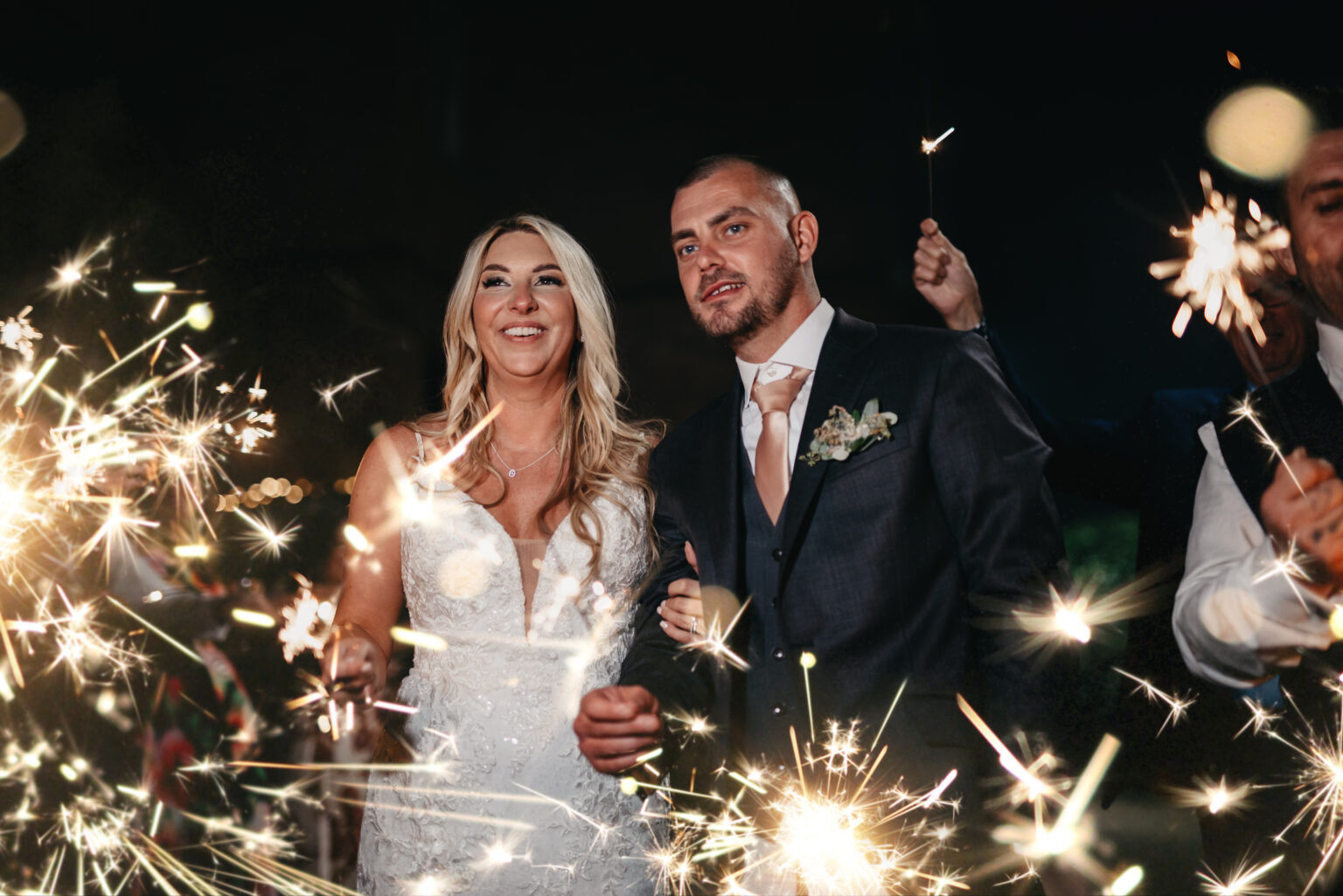 A bride in a white dress and a groom in a suit stand closely together, smiling as they hold lit sparklers. They are surrounded by friends, creating a festive and joyful atmosphere with more sparklers in the background.