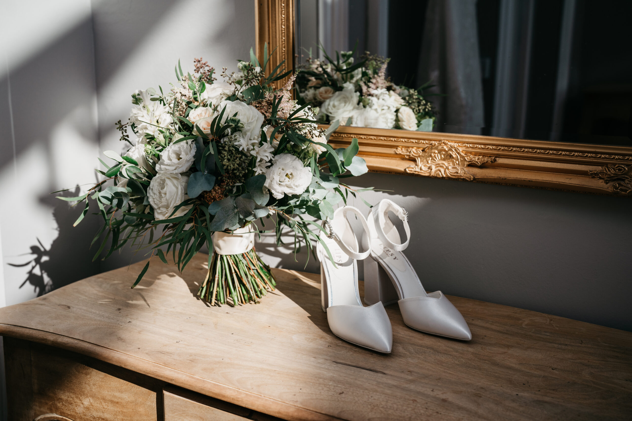 A bouquet of white flowers and greenery sits on a wooden dresser next to a pair of white heeled shoes with ankle straps. A large gold-framed mirror reflects both the bouquet and the shoes, with sunlight casting soft shadows.