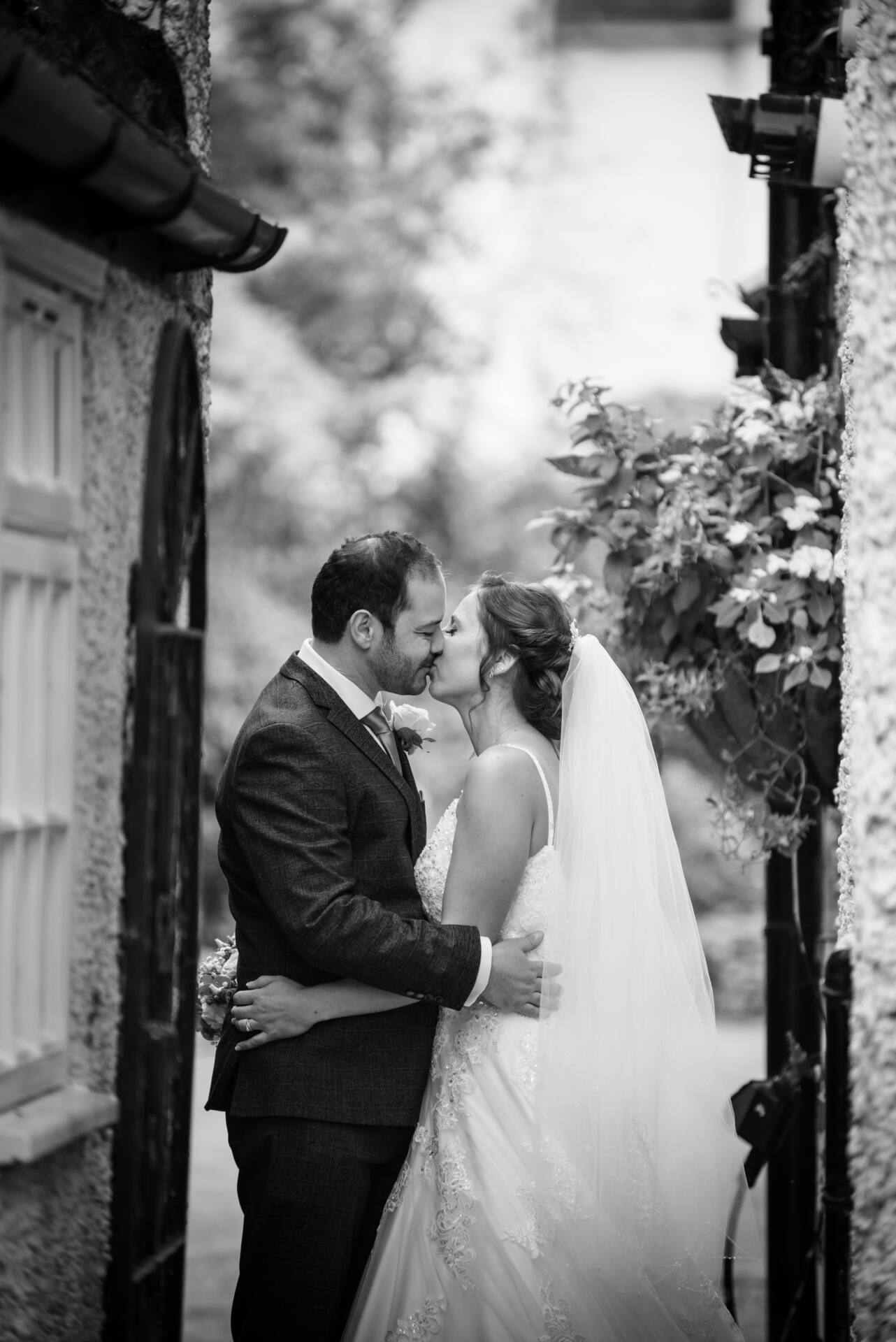 A bride and groom share a kiss in a narrow alleyway between two buildings. The bride wears a veil and lace gown, while the groom is in a dark suit. They are surrounded by greenery, creating a romantic atmosphere. The image is in black and white.