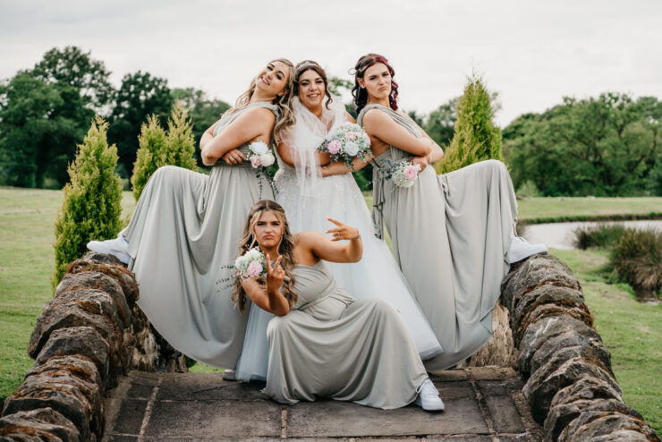 A bride in a white gown poses with three bridesmaids wearing grey dresses and white sneakers. They strike playful poses on a stone bridge, holding pink and white bouquets. Greenery and trees can be seen in the background.