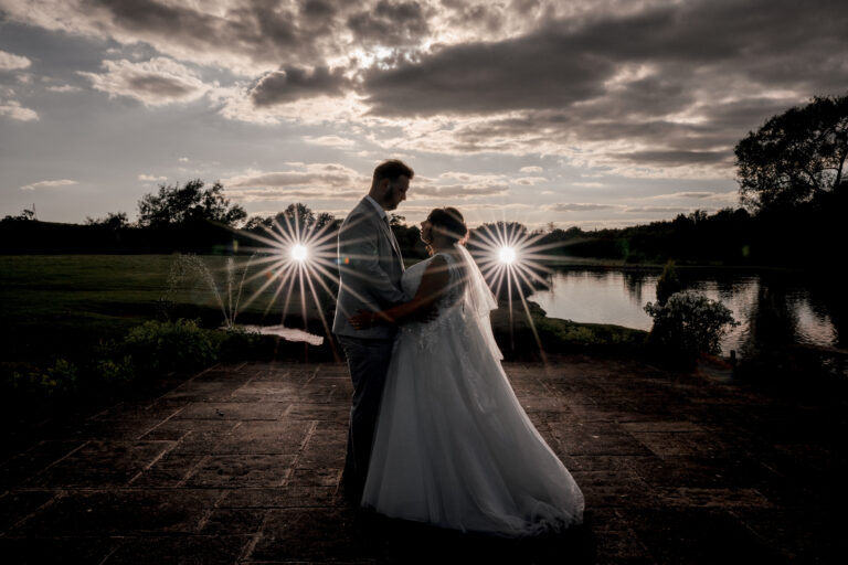 A bride and groom stand closely together, silhouetted against a dramatic sky with clouds. Two intense light flares frame them, creating a starburst effect. They are near a tranquil water body with trees and grass in the background.