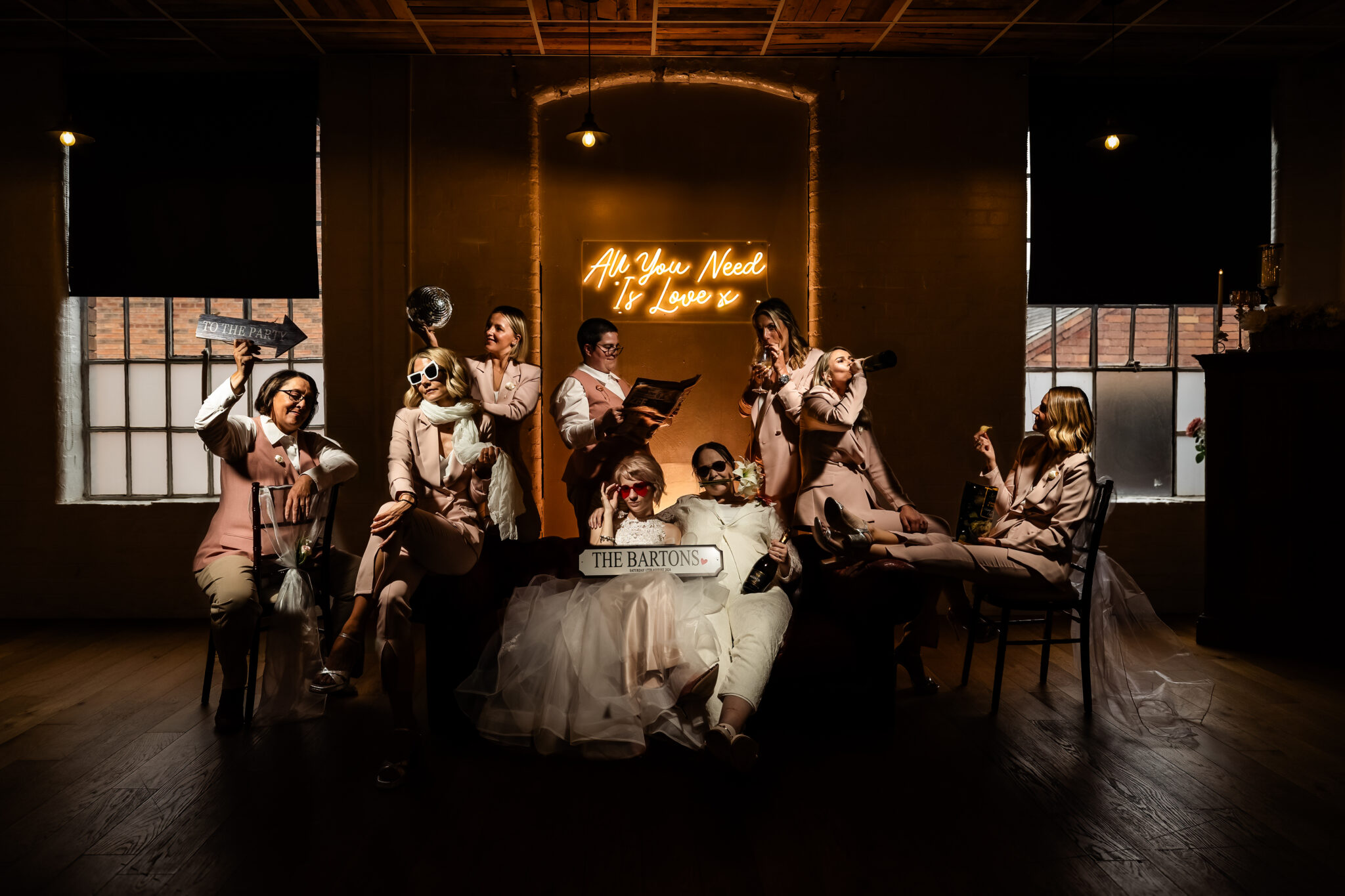 A group of people in formal attire and sunglasses are posing playfully in a dimly lit room. They hold props, including a bouquet, book, and signs. A neon sign in the background reads "All You Need Is Love.