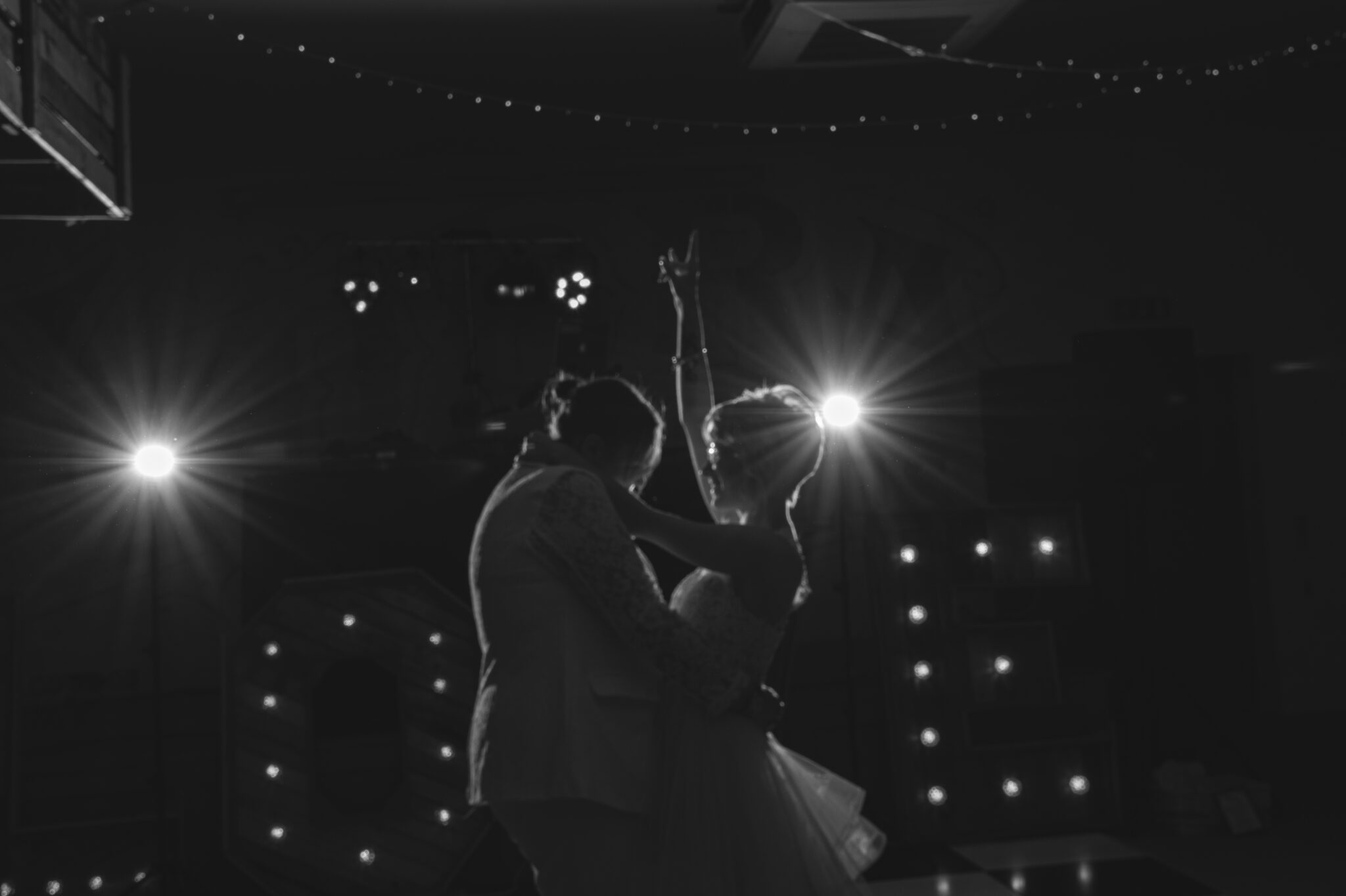 A couple shares a dance in a dimly lit room, backlit by soft fairy lights. The woman, wearing a wedding dress, raises one arm joyfully. The scene has a romantic and celebratory atmosphere.