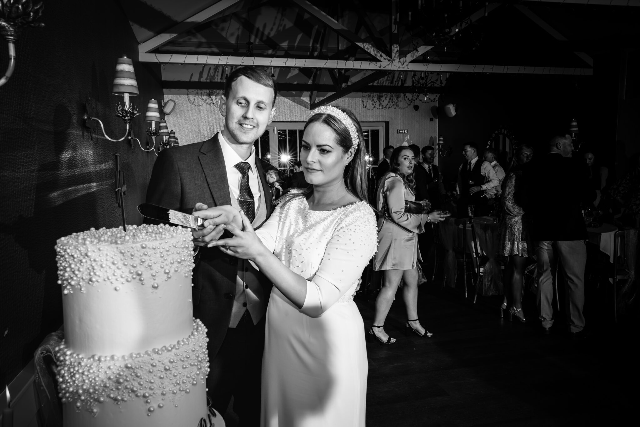 A bride and groom in formal attire smile as they cut a two-tiered wedding cake decorated with pearls. Guests are visible in the background, socializing in a dimly lit room. The scene is captured in black and white.