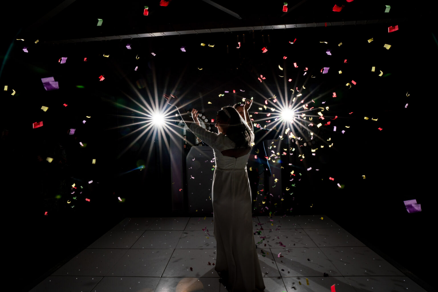 Silhouetted figure in a dress dances on a dimly lit floor, illuminated by bright stage lights. Colorful confetti floats around, adding a festive atmosphere.