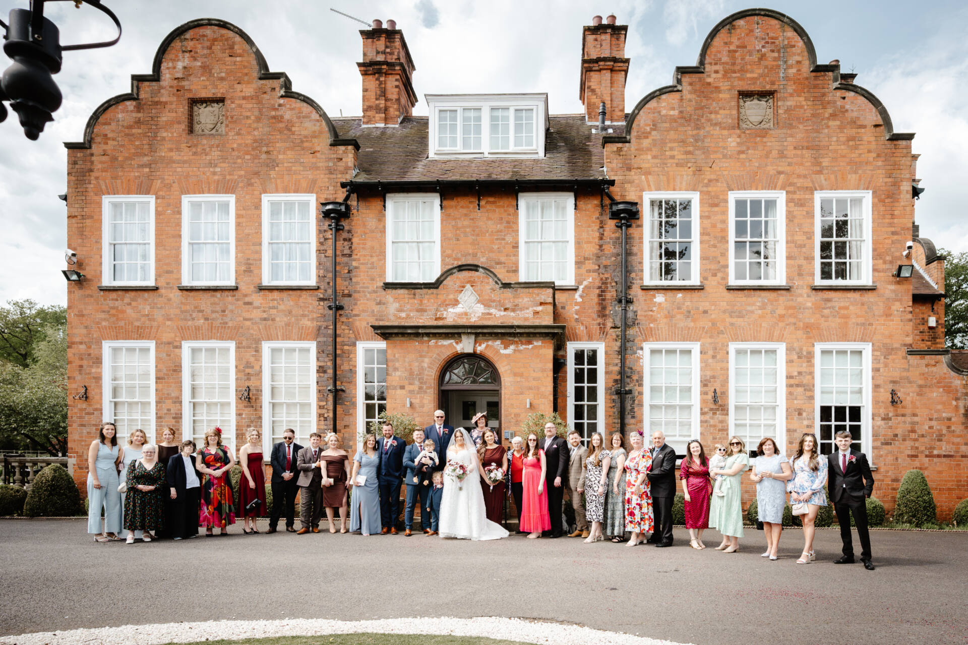 A large group of people dressed in formal attire pose for a photo in front of a grand red-brick building, beautifully captured by a Kelham wedding photographer, with white windows and decorative gables on a cloudy day.