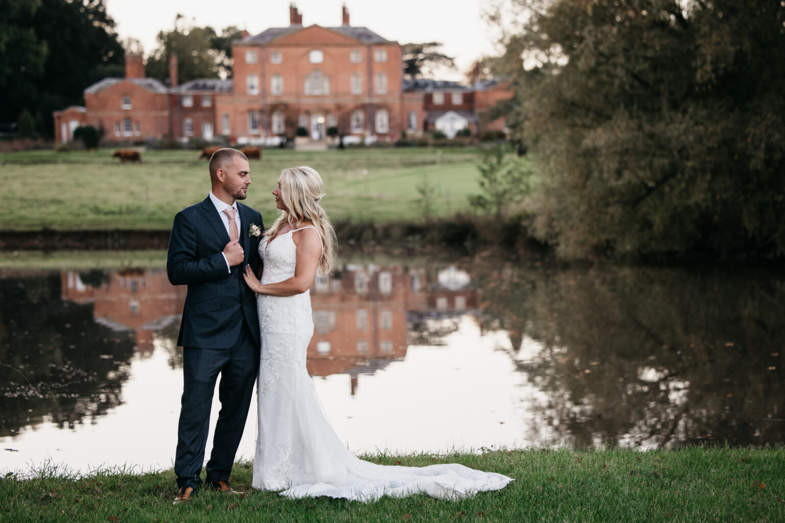 A bride and groom stand together by a calm lake at their Norwood Park wedding, gazing at one another, with a large elegant mansion and green lawns reflected in the water—captured by a skilled wedding photographer Nottinghamshire.